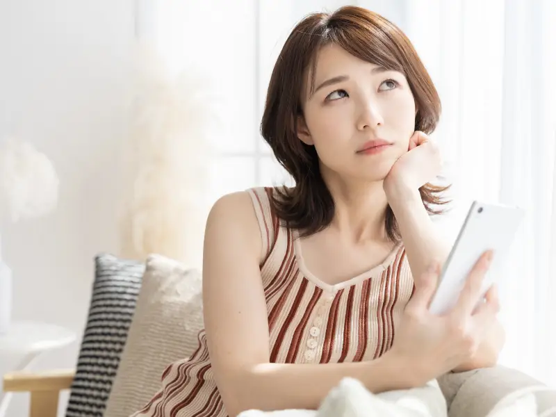 ソファーに腰を掛けてスマホを持ちながら悩んだ表情をしている女性